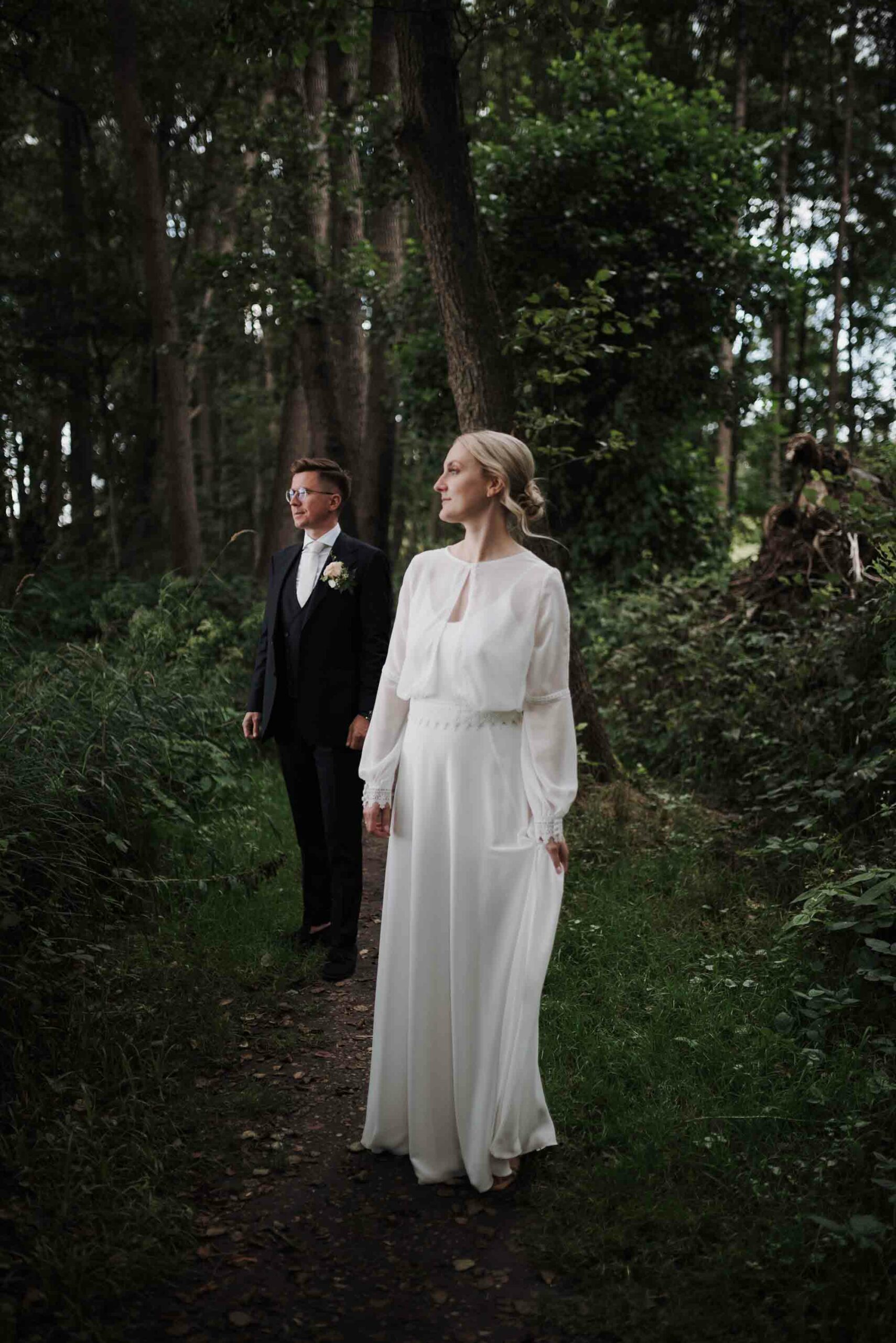 Ein elegantes Brautpaar im Wald nahe dem Seehotel Töpferhaus. Die Braut trägt ein fließendes, weißes Hochzeitskleid mit langen Ärmeln, und der Bräutigam ist in einem klassischen schwarzen Anzug gekleidet. Hochzeitsfotografin Schleswig Holstein hat diesen besonderen Moment festgehalten.