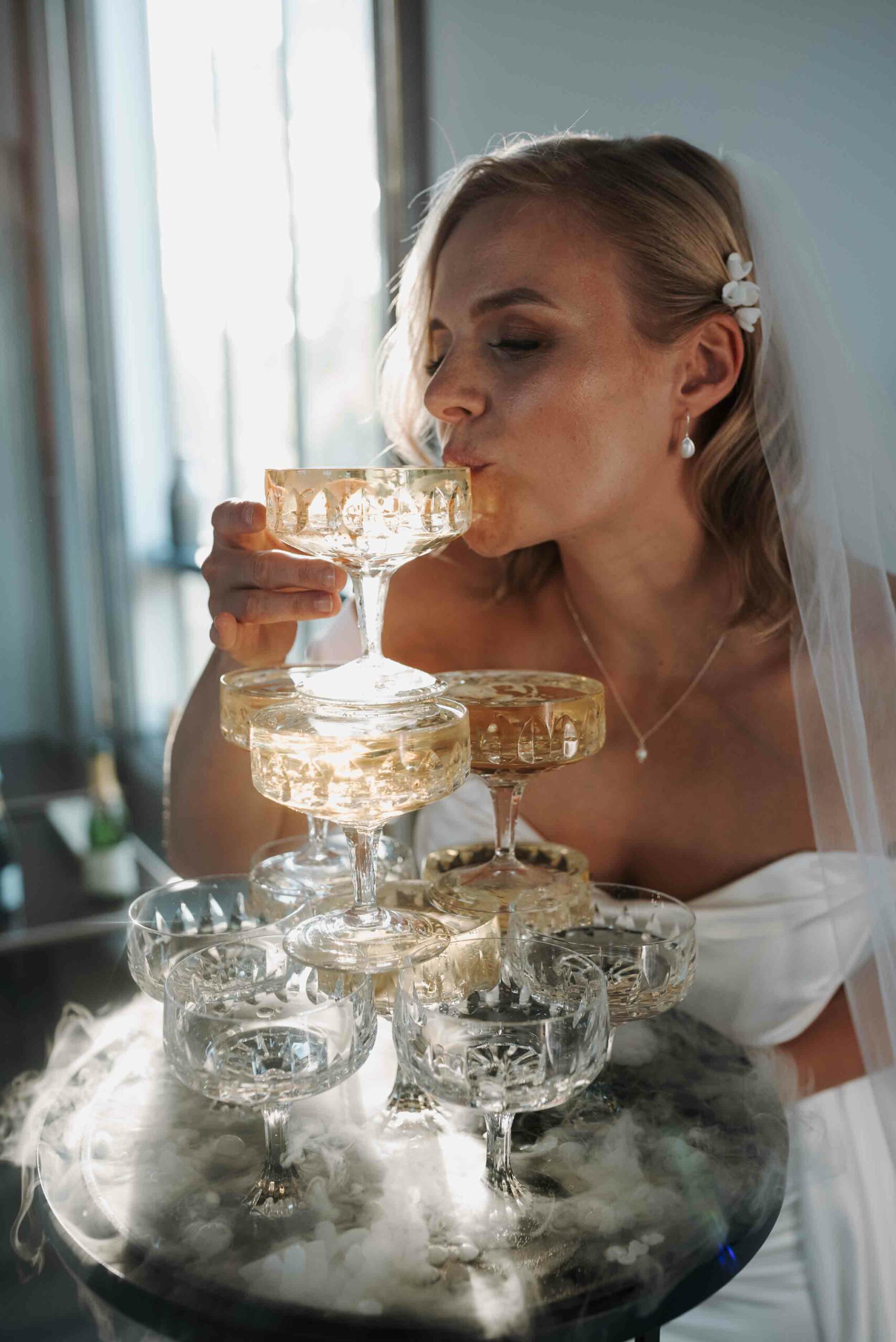 Eine Braut in einem weißen Kleid und Schleier genießt einen Drink aus einem eleganten Turm aus Champagnergläsern. Die gläsernen Kelche sind mit goldenem Sekt gefüllt und fangen das Licht der Umgebung ein, was dem Moment eine glamouröse Atmosphäre verleiht. Das Foto wurde aufgenommen auf einer Hochzeit in Hamburg.