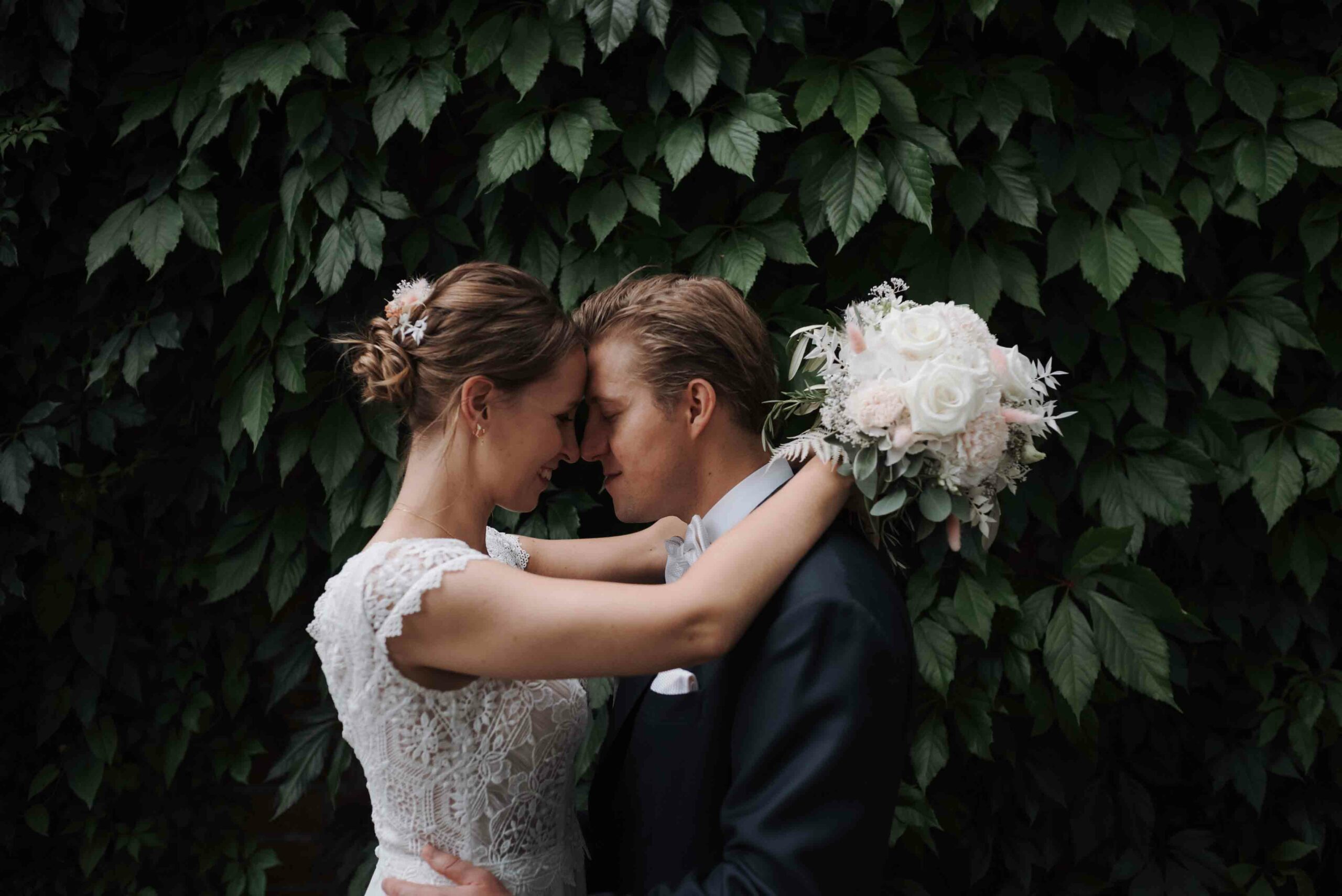 Ein Brautpaar in inniger Umarmung vor einer grünen Blätterwand an einem Ort nahe dem Elbstrand in Hamburg. Die Braut hält einen Strauß aus weißen Rosen und der Bräutigam trägt einen dunklen Anzug. Der Hochzeitsfotograf in Hamburg hat diesen romantischen Moment festgehalten.